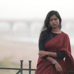 An elegant outdoor portrait of a woman in a red saree with a blurred bridge in the background.