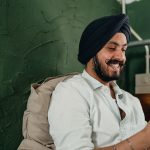 Young bearded Indian man in white shirt and dark turban leaning on pillow and browsing net on smartphone while spending time at home